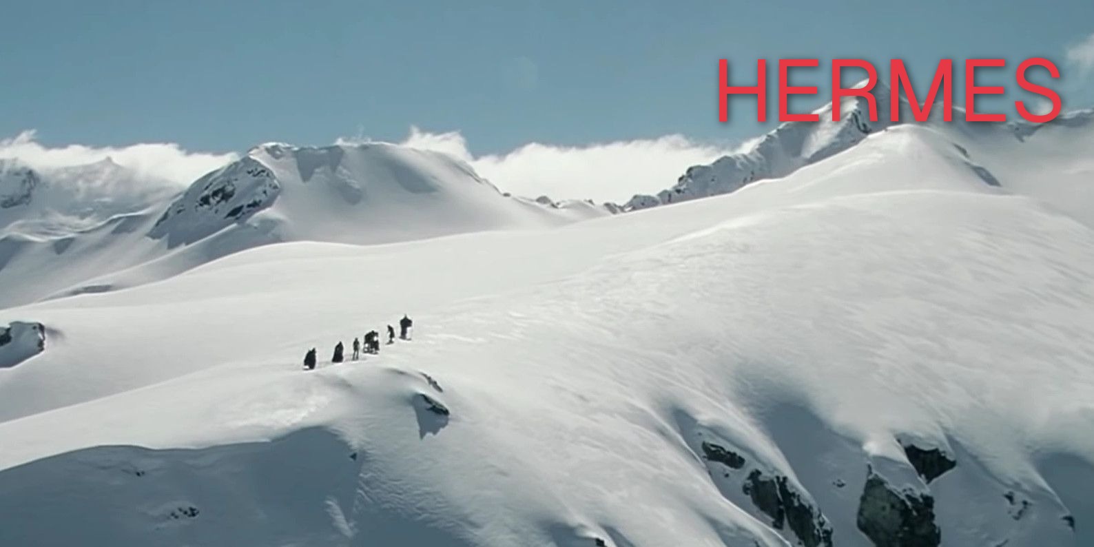 A film still from The Lord of the Rings: Fellowship of the Ring &mdash; the Fellowship as small figures crossing a vast snow ridge on Caradhras, with the word HERMES set in red Geist beside the distant peak.