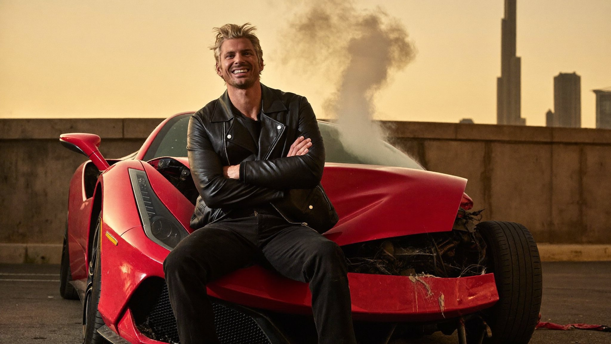 A grinning, leather-jacketed man sits utterly relaxed on the bonnet of a destroyed red Ferrari, crashed into a wall at golden hour with the Dubai skyline behind.
